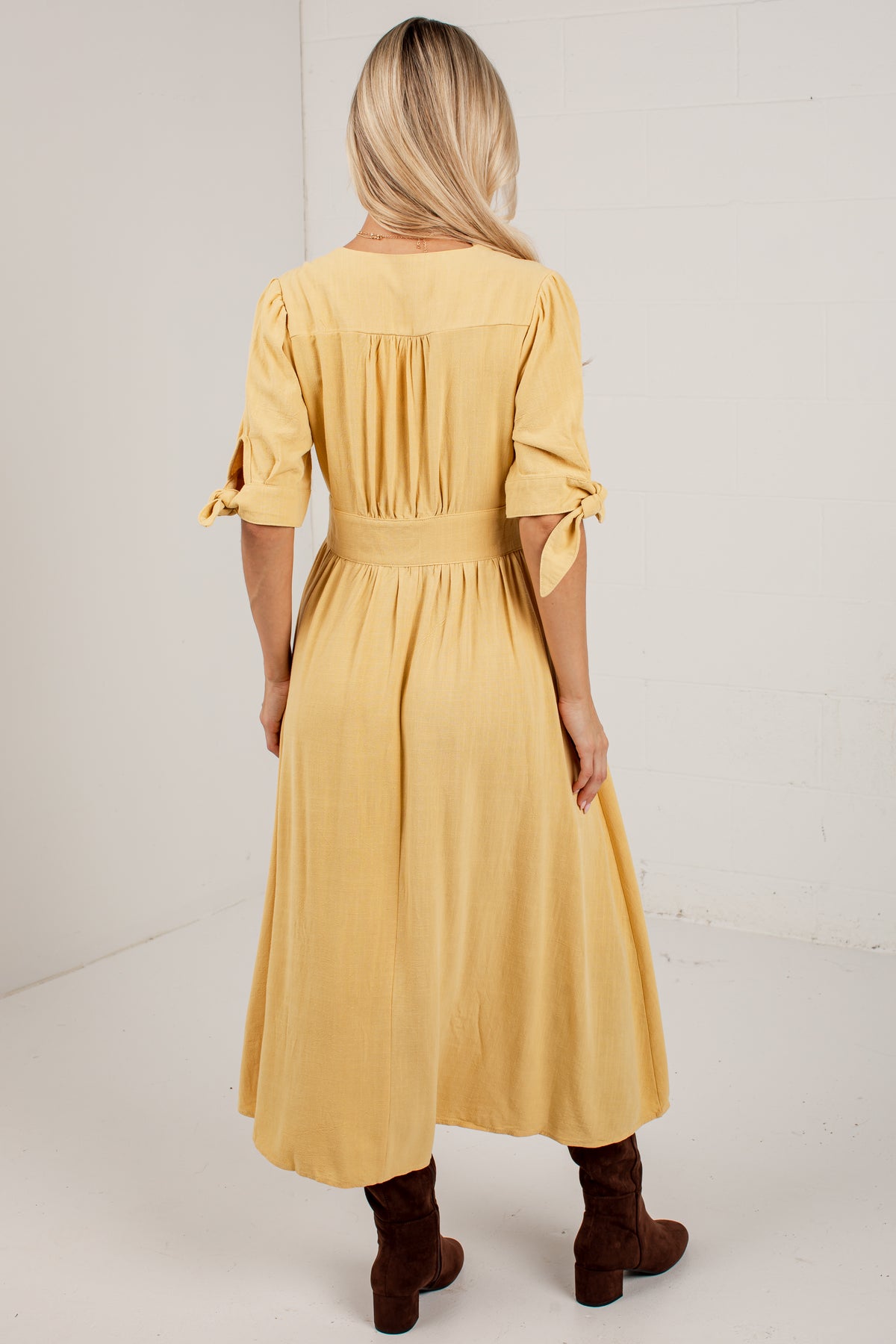 A woman viewed from behind wearing a mustard yellow midi dress with pleated back detailing, tie details on the sleeves, and brown ankle boots, set against a plain white studio background.