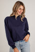 A woman wearing a navy crewneck sweatshirt and light blue jeans, standing against a solid light gray background.