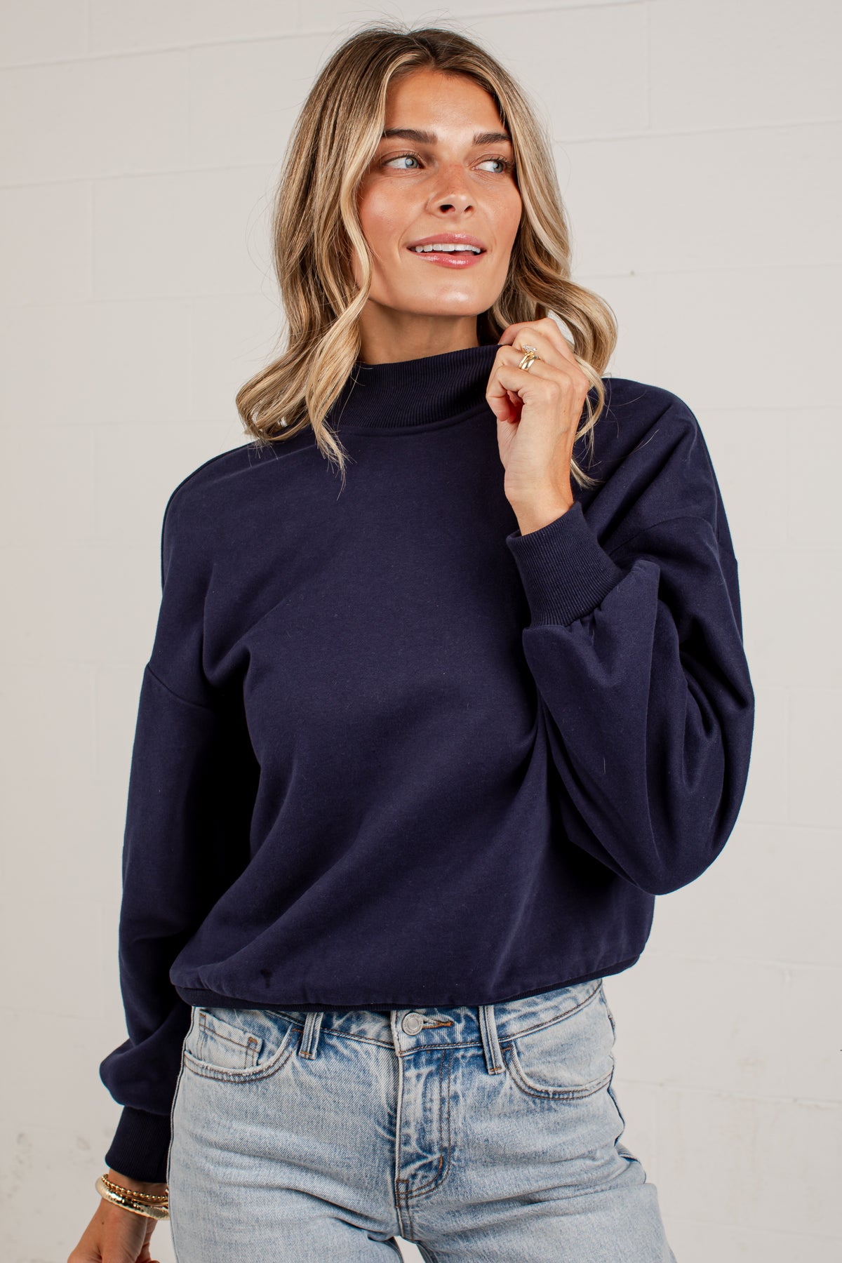 A woman wearing a navy crewneck sweatshirt and light blue jeans, standing against a solid light gray background.