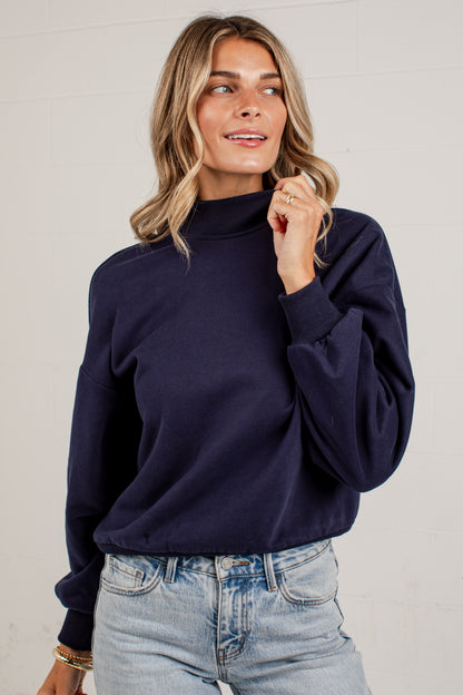 A woman wearing a navy crewneck sweatshirt and light blue jeans, standing against a solid light gray background.