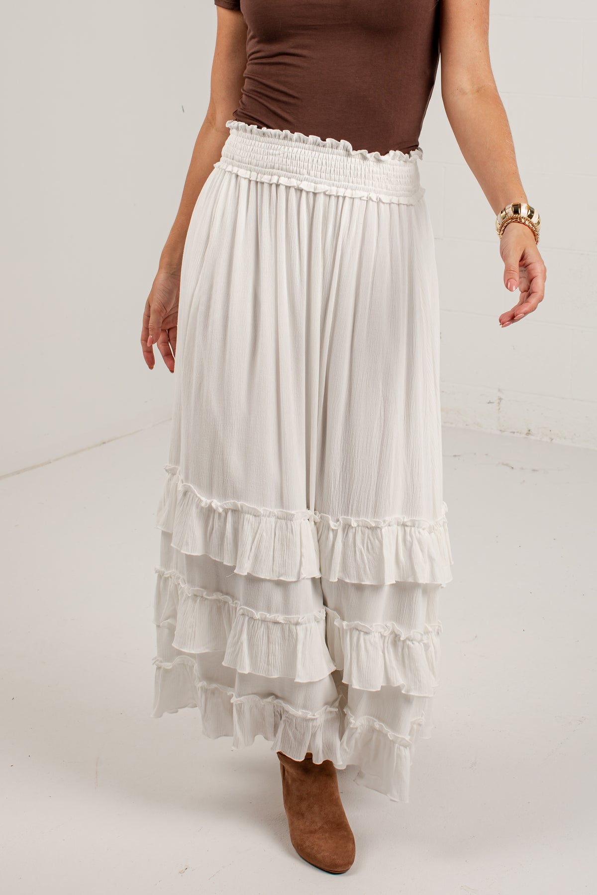 A model wearing a long white tiered maxi skirt with a smocked waistband, paired with a brown top and brown suede boots, shown from the chest down against a plain white studio background.