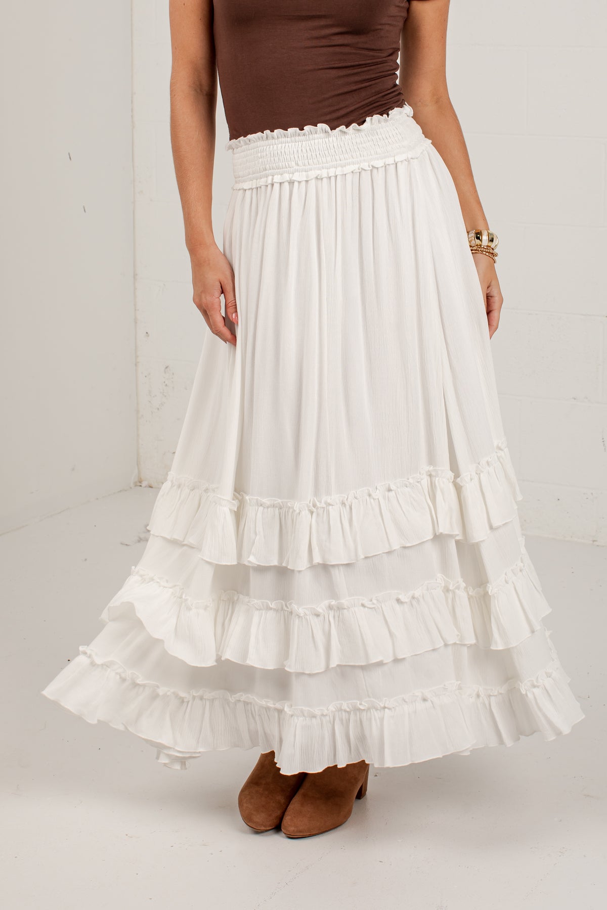 A model wearing a long white tiered maxi skirt with a smocked waistband, paired with a brown top and brown suede boots.