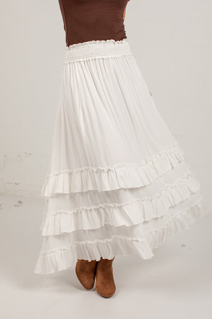A white tiered ruffle maxi skirt with a smocked elastic waistband, shown on a model wearing brown suede boots against a light off-white background.