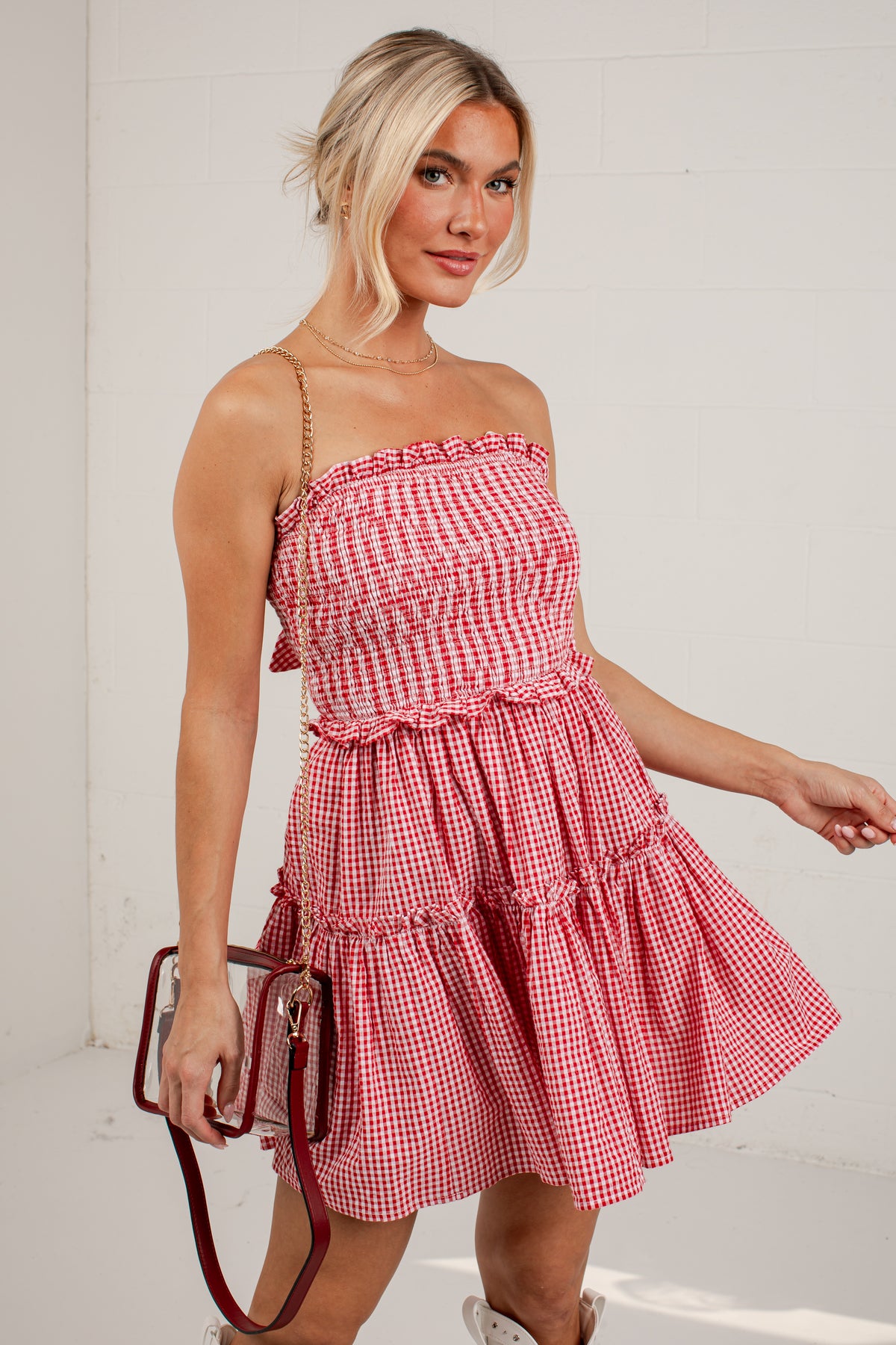 A woman modeling a red and white gingham tiered mini dress with a smocked bodice and thin straps, holding a small red handbag against a white studio background.