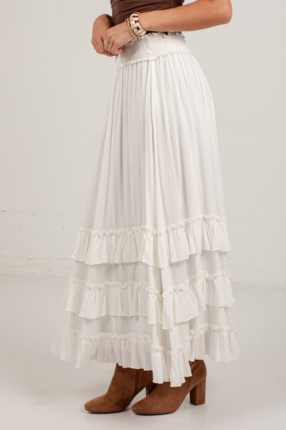 A white tiered maxi skirt with a smocked waistband, shown on a model wearing brown suede boots against a white brick wall.