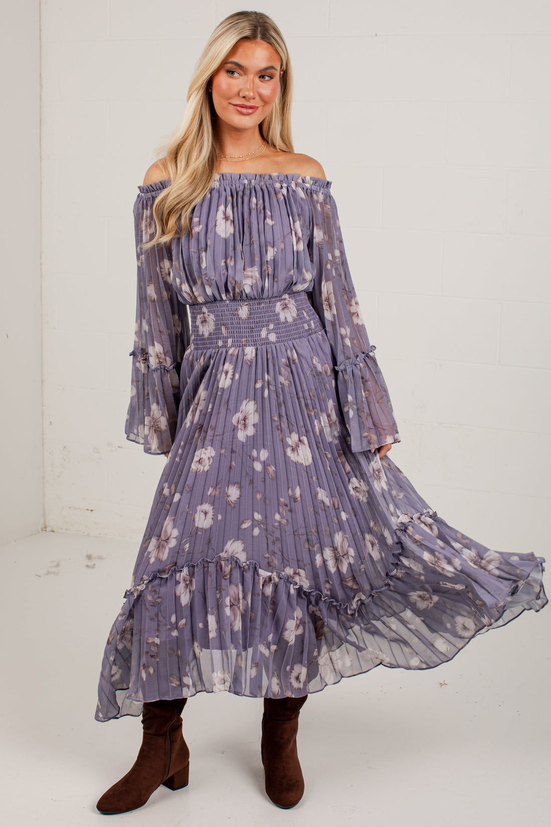 A woman modeling an off-the-shoulder midi dress in a dusty purple floral print, paired with brown suede ankle boots, standing against a plain white studio wall.