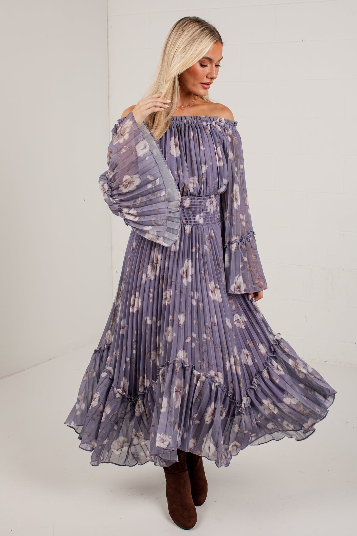 A woman modeling an off-the-shoulder, long-sleeved maxi dress with a purple and white floral print, paired with brown boots, in a bright studio setting.