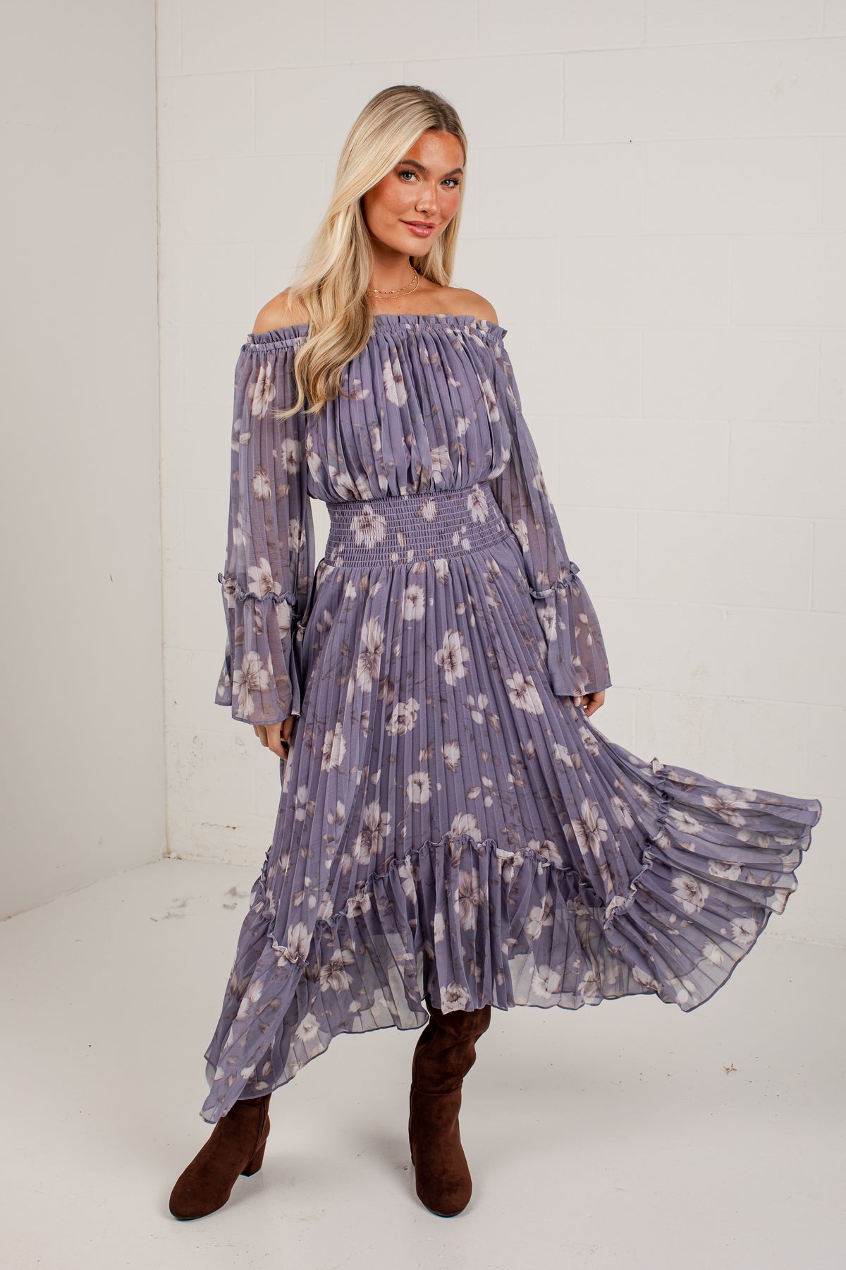 A woman modeling a long off-the-shoulder midi dress in a dusty lavender-purple color with a white floral pattern, paired with brown suede boots, standing against a white studio wall.