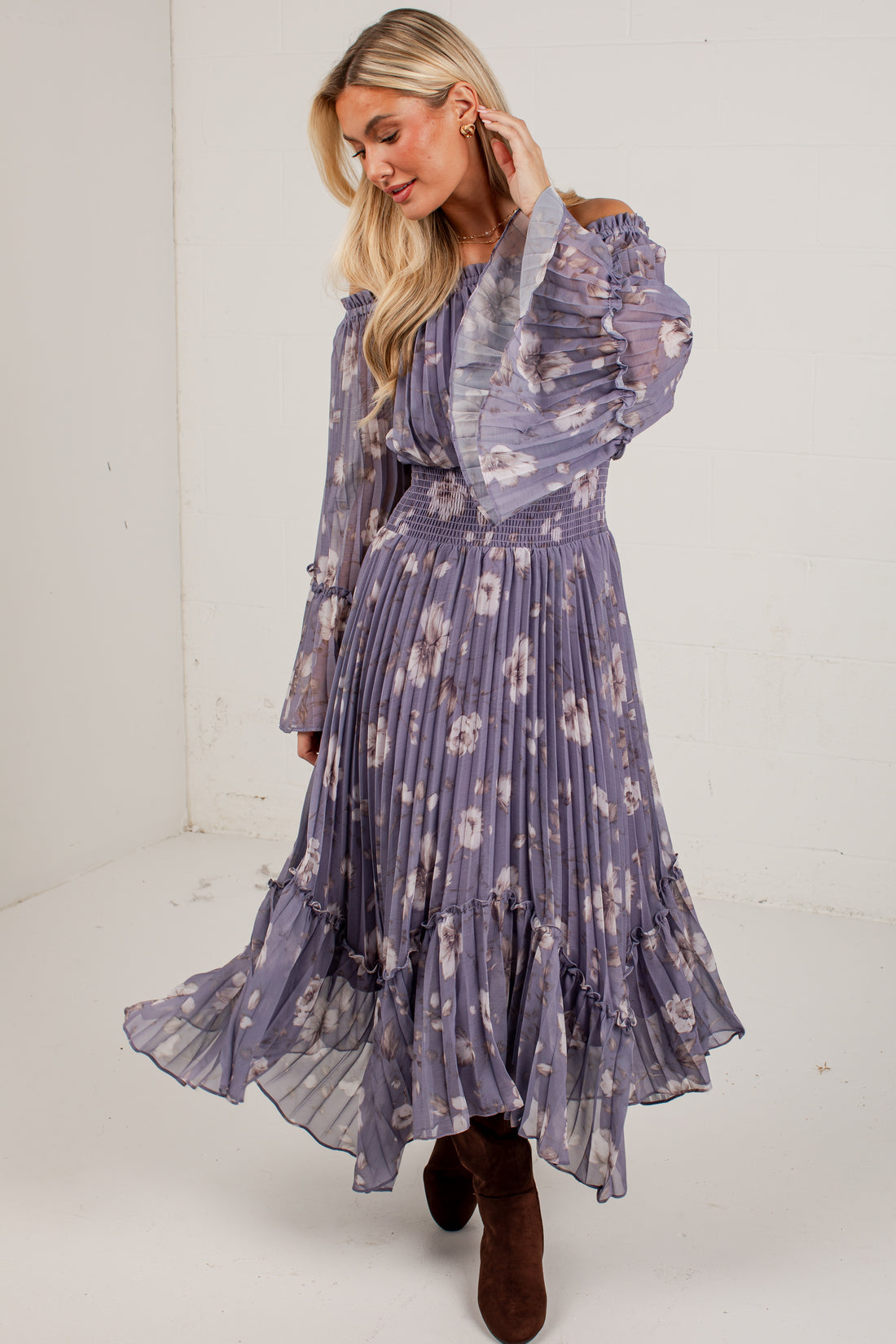 A woman modeling a long-sleeve midi dress with a purple and white floral print, featuring a pleated skirt and ruffled sleeves, paired with brown suede boots in a white studio setting.