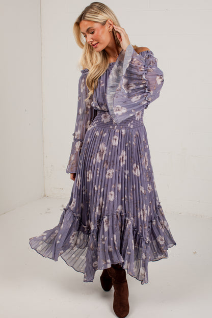 A woman modeling a long-sleeve midi dress with a purple and white floral print, featuring a pleated skirt and ruffled sleeves, paired with brown suede boots in a white studio setting.