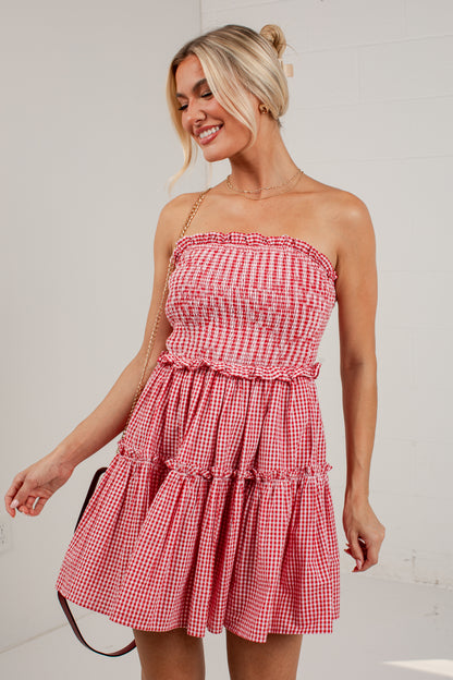 A woman wearing a red and white gingham strapless tiered dress, holding a black handbag against a white wall.