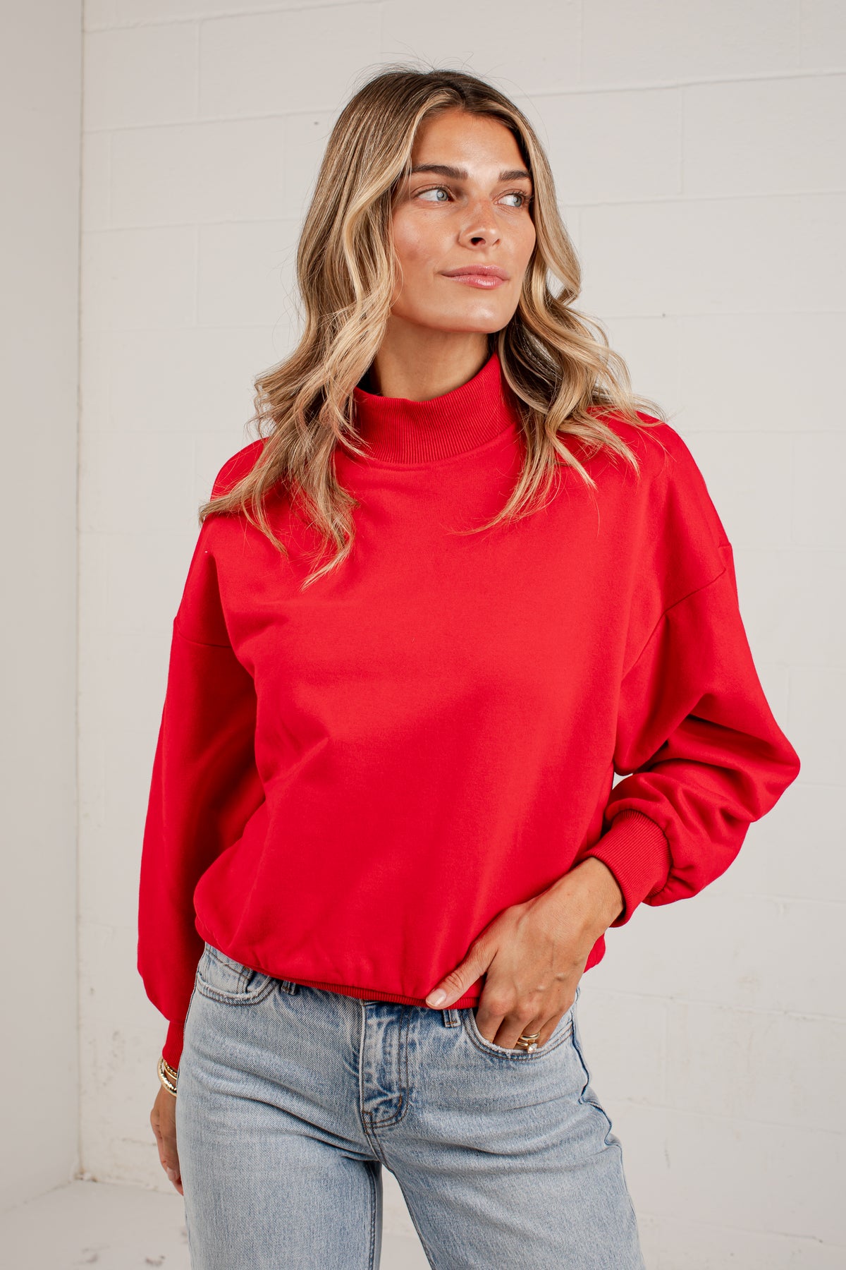 A woman wearing a red mockneck sweatshirt and light blue jeans, standing against a solid light gray background.