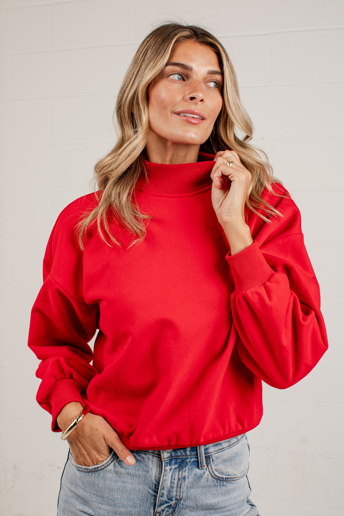 A woman wearing a red mockneck sweatshirt and light blue jeans, standing against a solid light gray background.