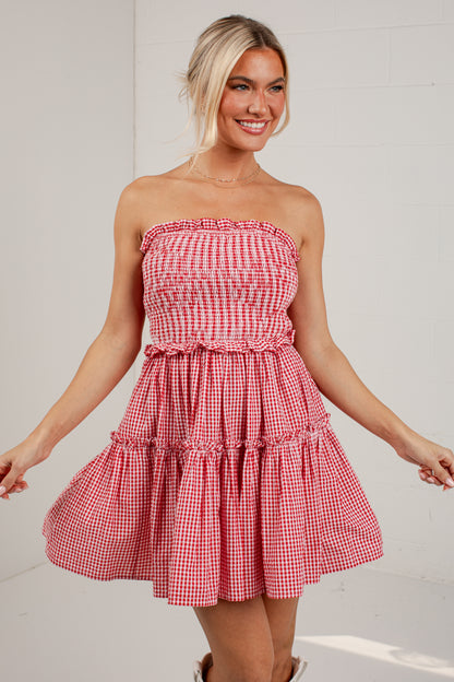 A woman modeling a strapless red and white gingham tiered dress against a white brick wall background.