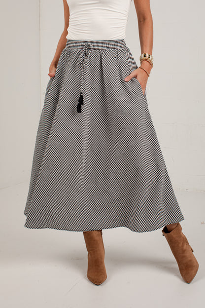 A woman wearing a high-waisted, A-line midi skirt with a small black and white micro-pattern, paired with a white top and brown suede ankle boots.