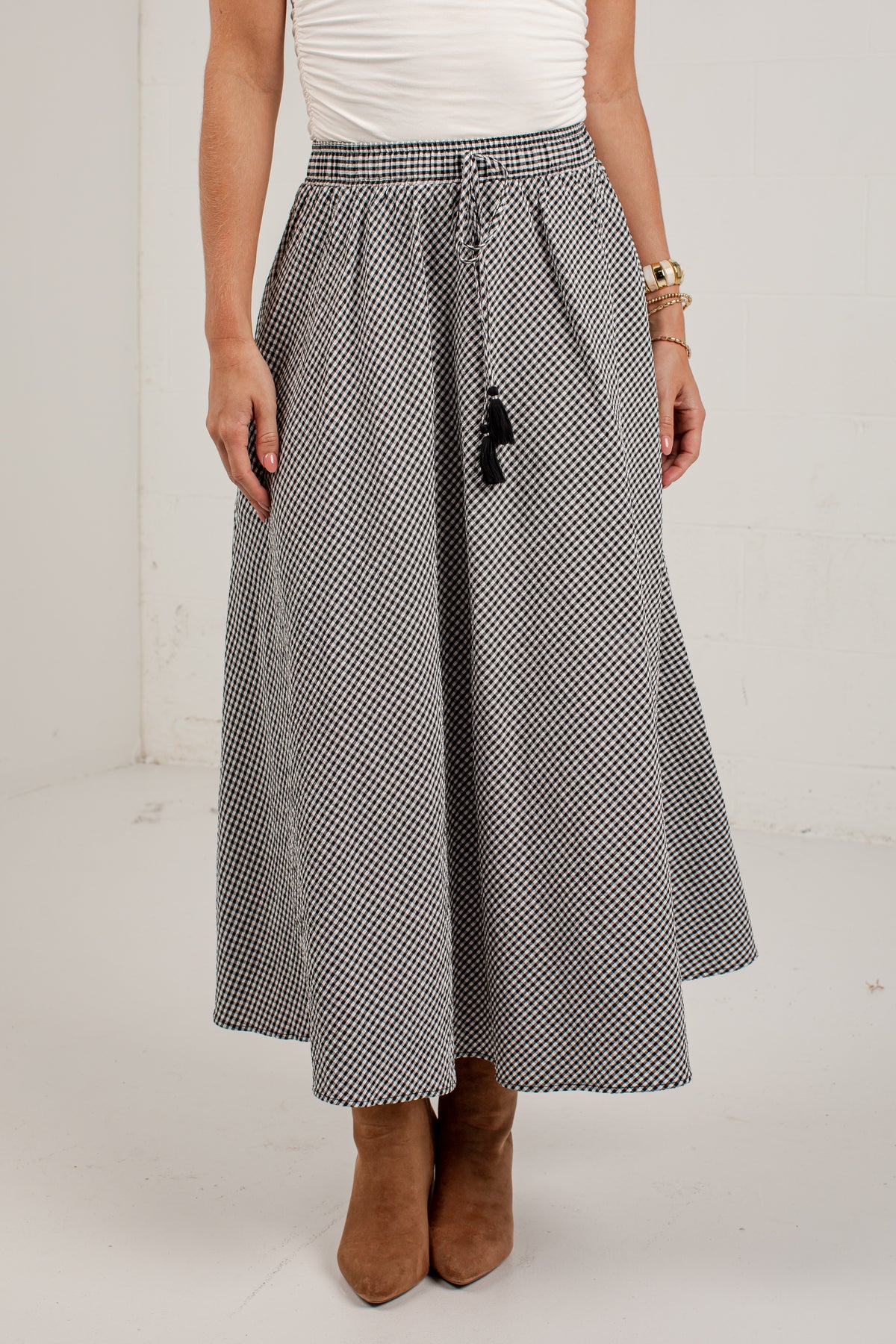 A woman wearing a black and white micro-checkered midi skirt with a drawstring waist, paired with a white top and brown suede ankle boots.