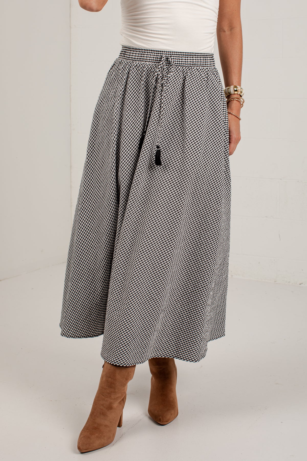 A model wearing a black and white micro-checkered midi skirt with a drawstring waist, paired with a white top and brown suede ankle boots.