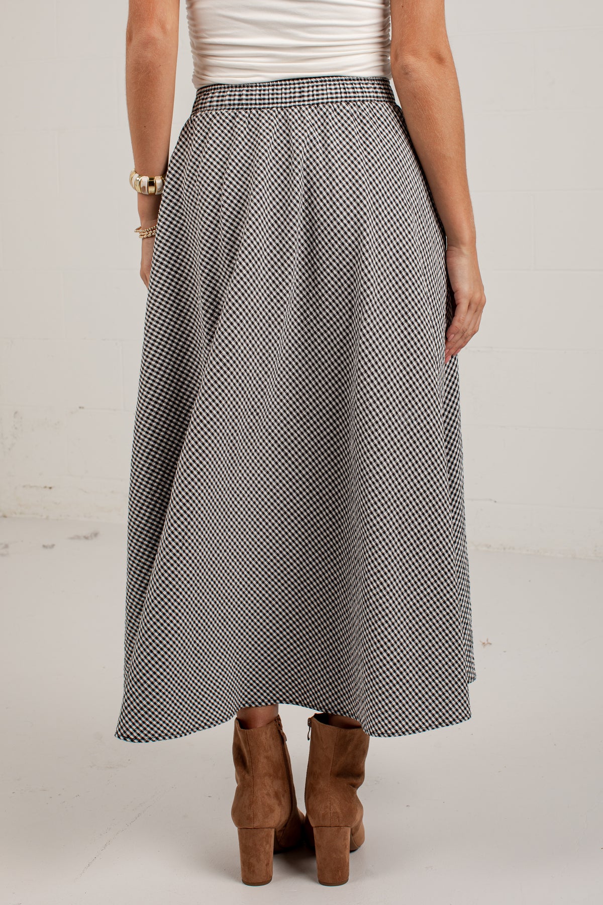 A woman wearing a black and white micro-checkered midi skirt, paired with a white ribbed top and brown suede ankle boots, shown from the back against a plain off-white studio background.