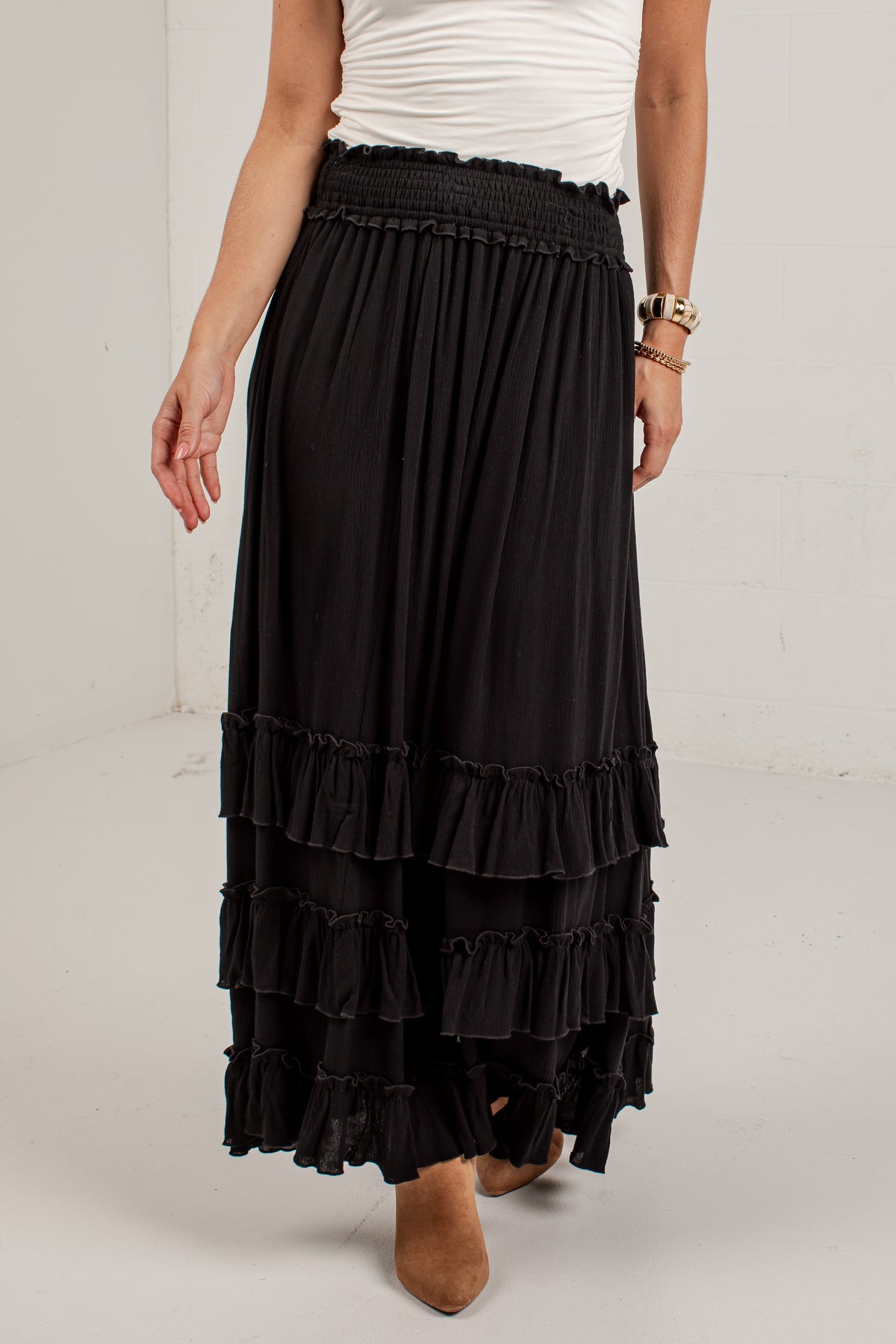 A black tiered maxi skirt with a smocked waistband, shown on a model wearing a white top and brown boots against a white studio background.