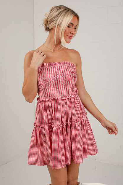 A woman modeling a red and white gingham strapless mini dress with a smocked bodice and tiered skirt, standing against a white brick wall.