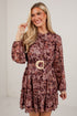 A woman modeling a long-sleeve mini dress with a brown and burgundy abstract print, featuring a matching belt with a gold-tone buckle, set against a white brick wall.