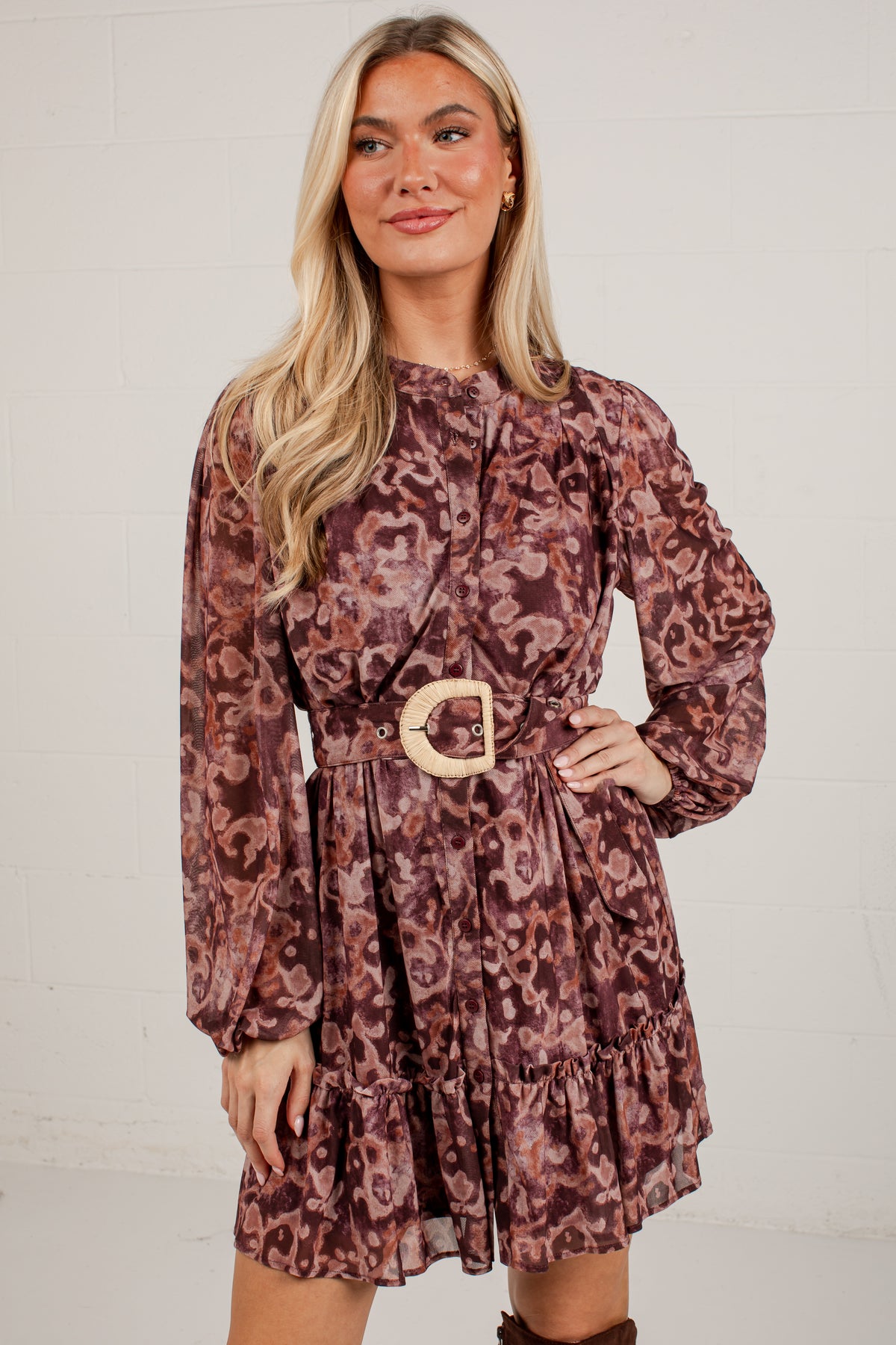 A woman modeling a long-sleeve mini dress with a brown and burgundy abstract print, featuring a matching belt with a large round gold buckle, set against a white brick wall.