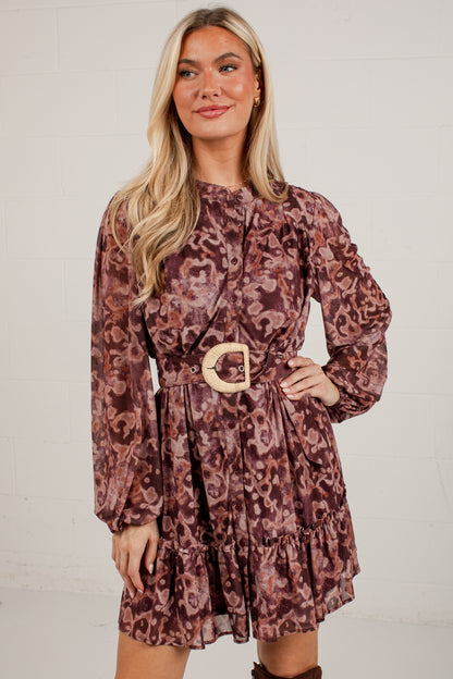 A woman modeling a long-sleeve mini dress with a brown and burgundy abstract print, featuring a matching belt with a large round gold buckle, set against a white brick wall.