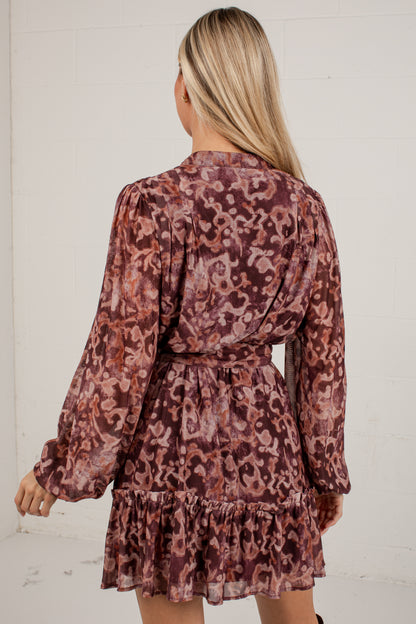 A woman viewed from the back wearing a long-sleeve mini dress with a dusty rose and brown abstract floral pattern, set against a white brick wall.