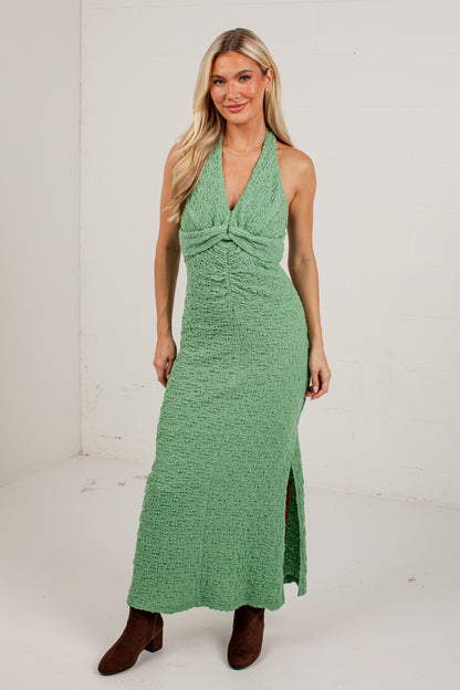 A woman modeling a sleeveless, halter-neck, floor-length green dress with a textured knit fabric, a twisted front detail, and a side slit, paired with brown ankle boots against a white studio background.