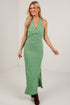 A woman modeling a sleeveless, halter-neck, floor-length green dress with a textured knit fabric, a twisted detail at the bust, and a side slit, paired with brown ankle boots against a white studio background.