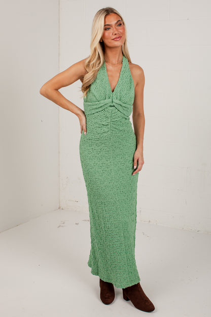 A woman modeling a sleeveless, halter-neck, floor-length green dress with a textured knit fabric, standing against a white studio wall.