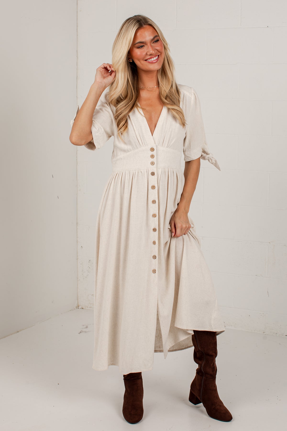 A woman modeling a cream-colored midi dress with a button-down front and tie details on the sleeves, paired with brown boots, standing in a white studio corner.