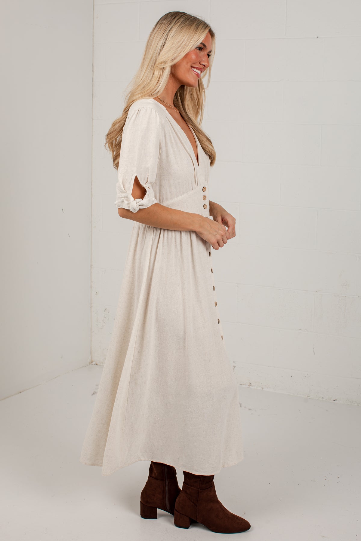 A woman modeling a cream-colored midi dress with elbow-length sleeves featuring cutouts, a V-neckline, and a button-down front, paired with brown ankle boots, set against a white brick wall background.