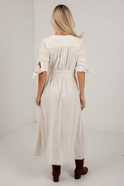 A woman viewed from behind wearing a cream-colored midi dress with three-quarter sleeves, tie details at the cuffs, and a gathered waist, paired with brown ankle boots in a white studio setting.