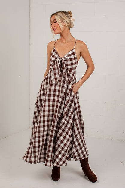 A woman modeling a brown and white gingham maxi dress with spaghetti straps, a front tie detail, and side pockets, paired with brown boots, set against a white brick wall background.