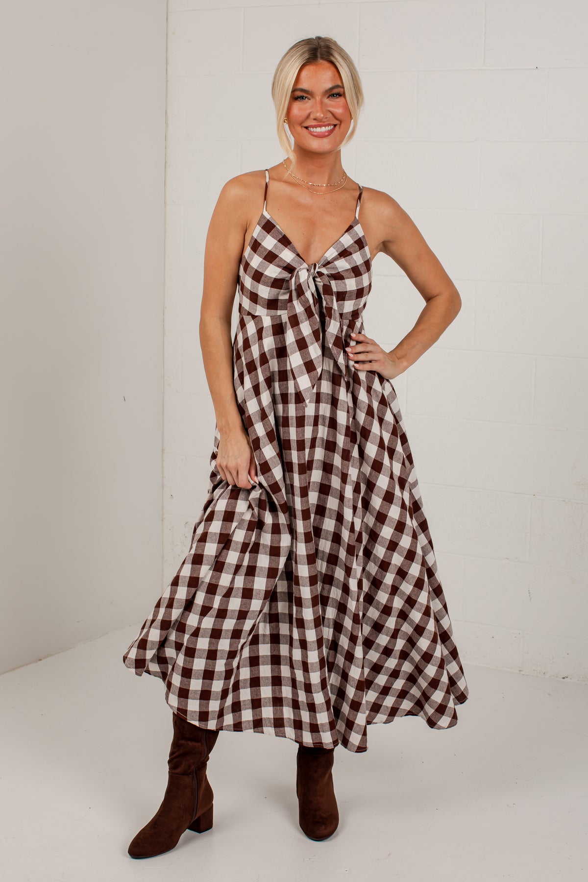 A woman modeling a brown and white gingham midi dress with spaghetti straps and a tie-front detail, paired with brown suede boots, set against a white brick wall background.