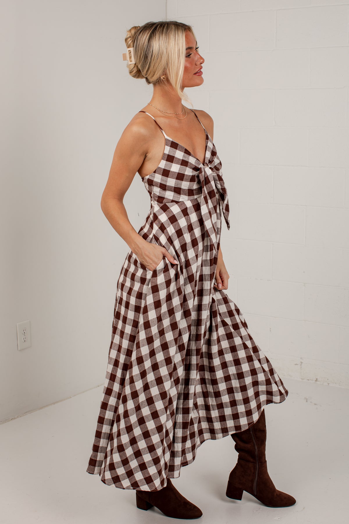A woman modeling a brown and white gingham wrap maxi dress with thin straps, side pockets, and a knotted bow detail at the bust, paired with brown knee-high boots against a white studio wall.