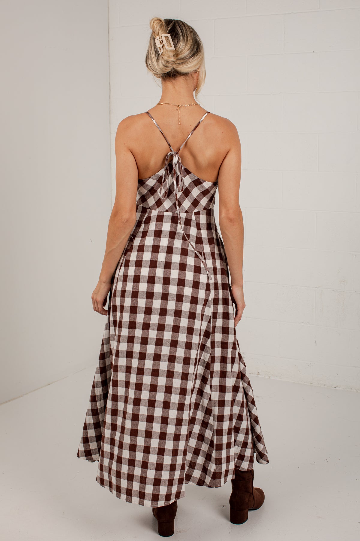 A woman viewed from behind wearing a brown and white gingham maxi dress with thin straps and a tie at the back, paired with brown boots, against a white studio background.
