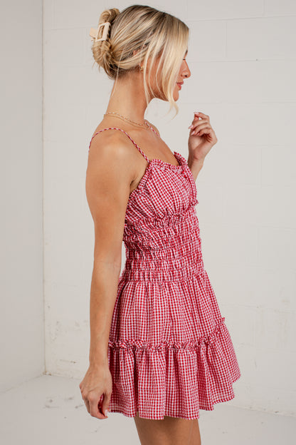 A woman wearing a red and white gingham tiered mini dress with thin straps, shown from a side profile against a white studio wall.