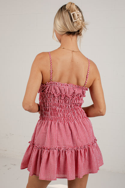 A woman shown from the back wearing a red and white gingham mini dress with thin spaghetti straps, a smocked bodice, and a tiered ruffle skirt, set against a white paneled wall.