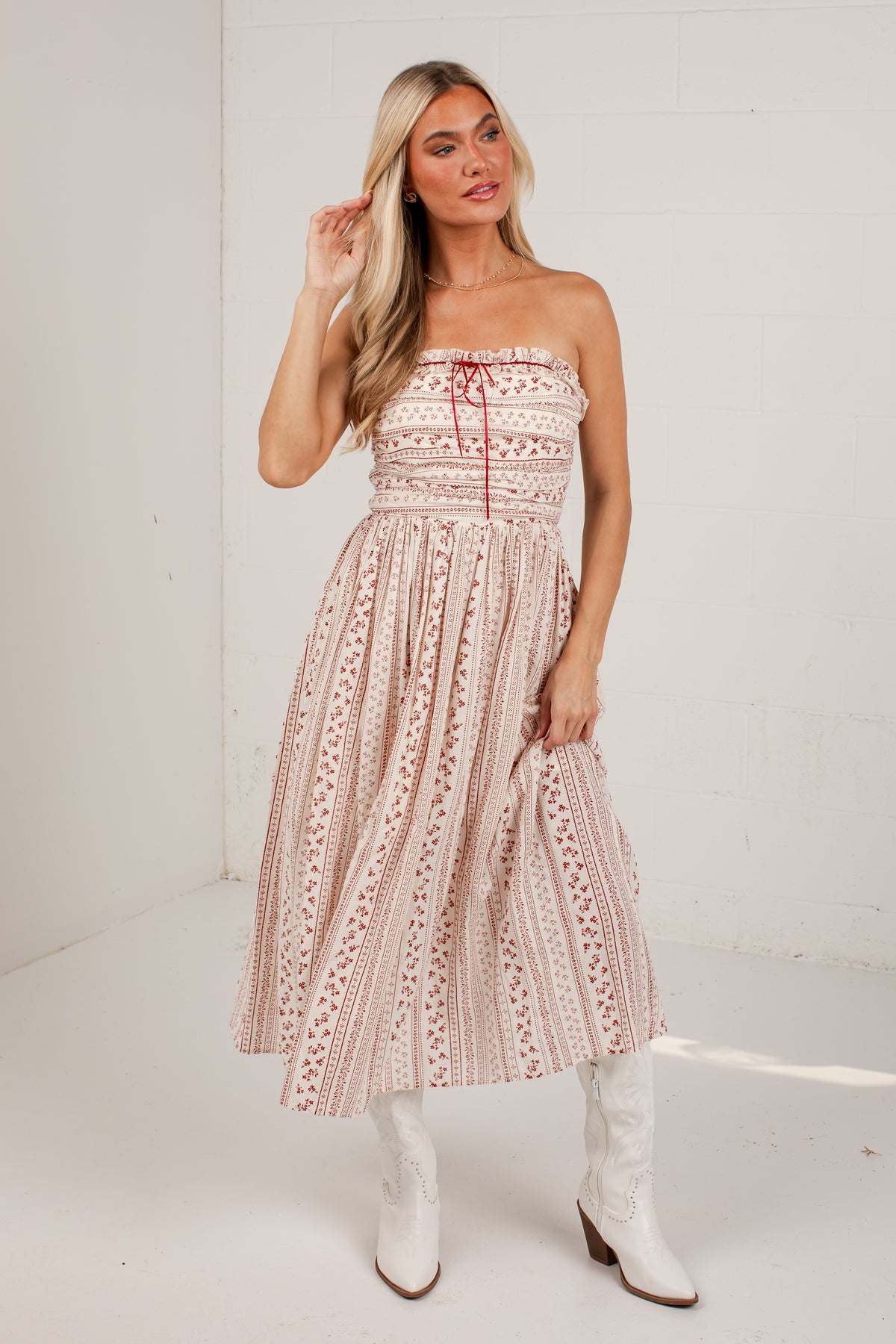 A woman modeling a strapless midi dress with a pink and beige bohemian pattern, paired with white cowboy boots, standing against a white brick wall.