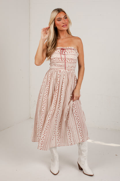 A woman modeling a strapless midi dress with a pink and beige bohemian pattern, paired with white cowboy boots, standing against a white brick wall.