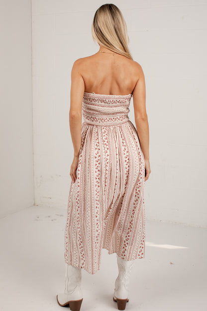 A woman shown from the back wearing a strapless midi dress with a smocked bodice and a pleated skirt, featuring a vertical floral pattern in dusty pink and white. She is wearing white cowboy boots in a bright, white-walled room.