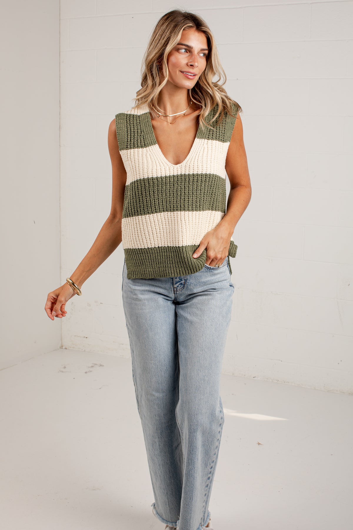 A woman modeling a sleeveless ribbed knit vest with horizontal olive green and cream stripes, paired with light-wash blue jeans, in a bright white studio setting.