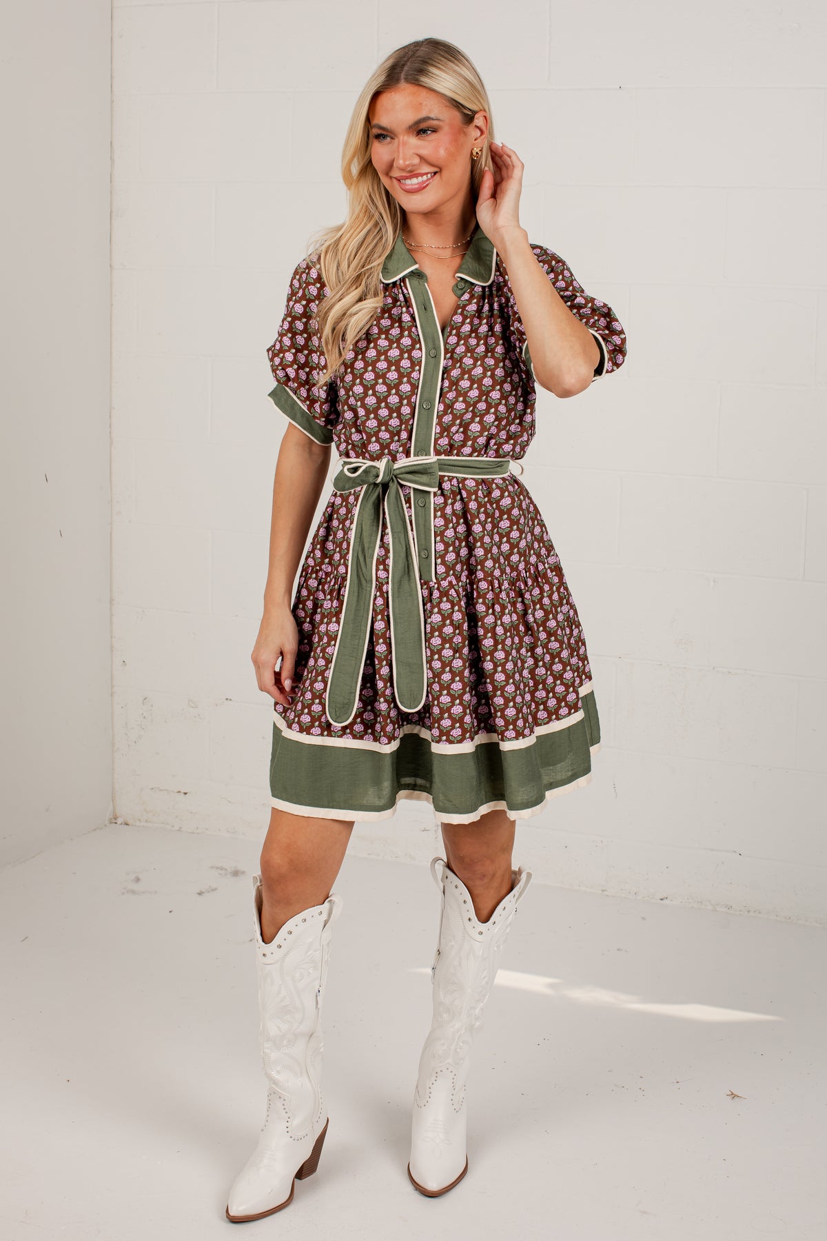 A woman modeling a short-sleeve brown and white floral print dress with sage green trim and a matching tie belt, paired with white cowboy boots, in a bright studio setting.