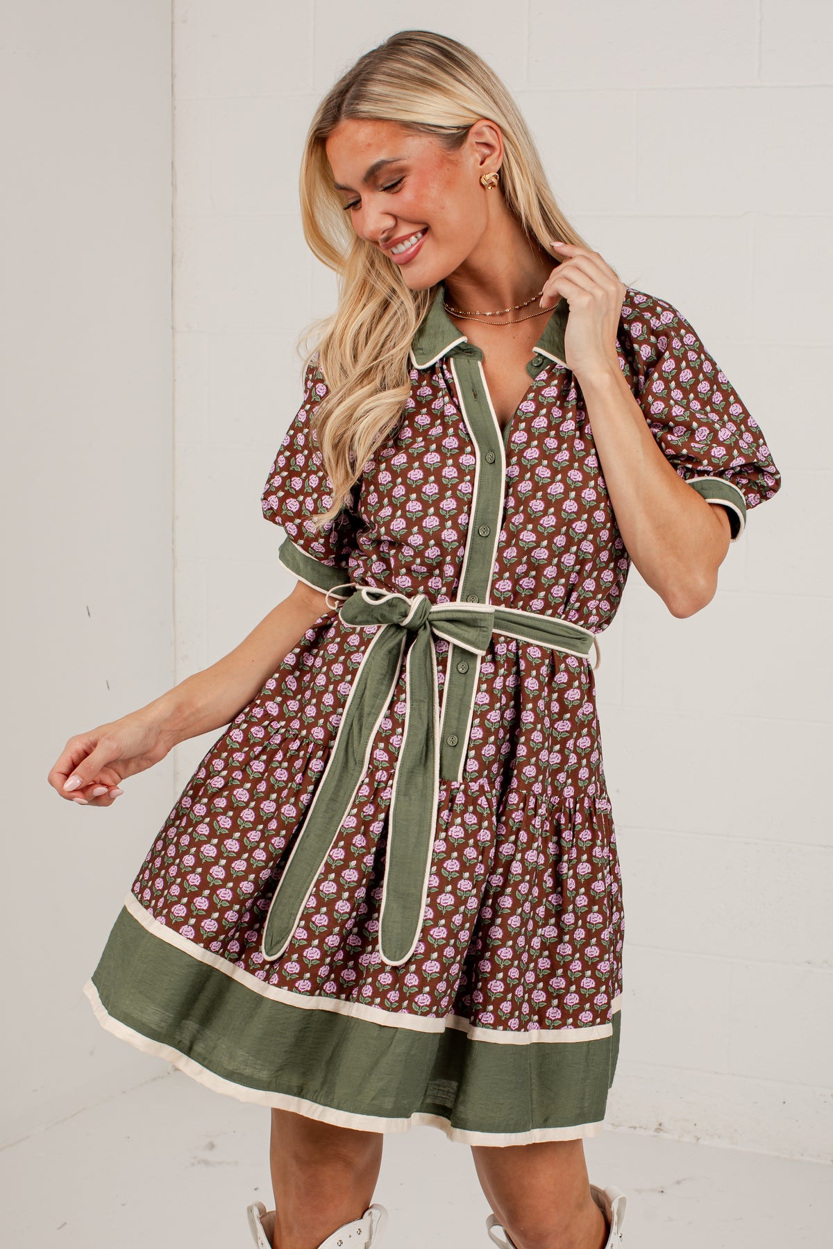 A woman modeling a short-sleeve button-front dress with a brown and beige floral print, olive green trim, and a matching green tie belt, set against a plain white wall.