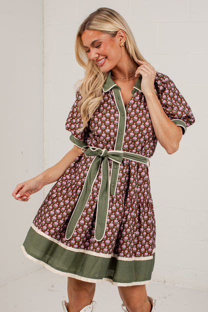 A woman modeling a short-sleeve button-front dress with a brown and beige floral print, olive green trim, and a matching green tie belt, set against a plain white wall.