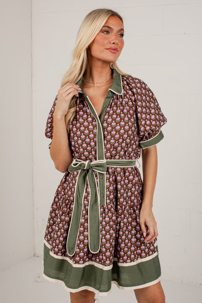A woman modeling a short-sleeve button-front dress with an allover floral print in brown, burgundy, and cream, with sage green trim and a matching tie belt.