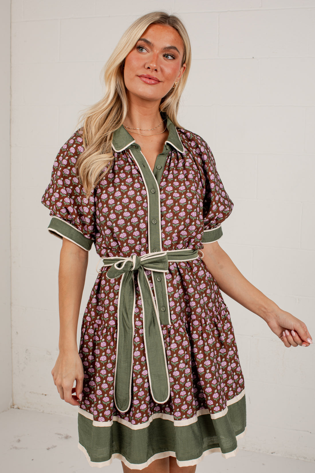 A woman modeling a short-sleeve button-front dress with an allover floral print in brown, green, and cream, featuring solid green trim, a matching green tie belt, and a green ruffled hem, set against a white brick wall.