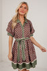 A woman modeling a short-sleeve button-front dress with an allover floral print in brown, green, and cream, featuring solid green trim, a matching green tie belt, and a green ruffled hem, set against a white brick wall.